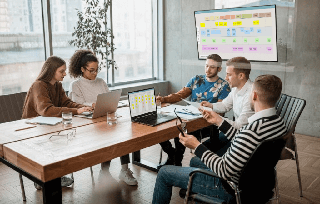 Five colleagues are in a modern meeting room collaborating, with two using laptops and others discussing notes around a wooden table, a large screen with colorful charts is visible on the wall.